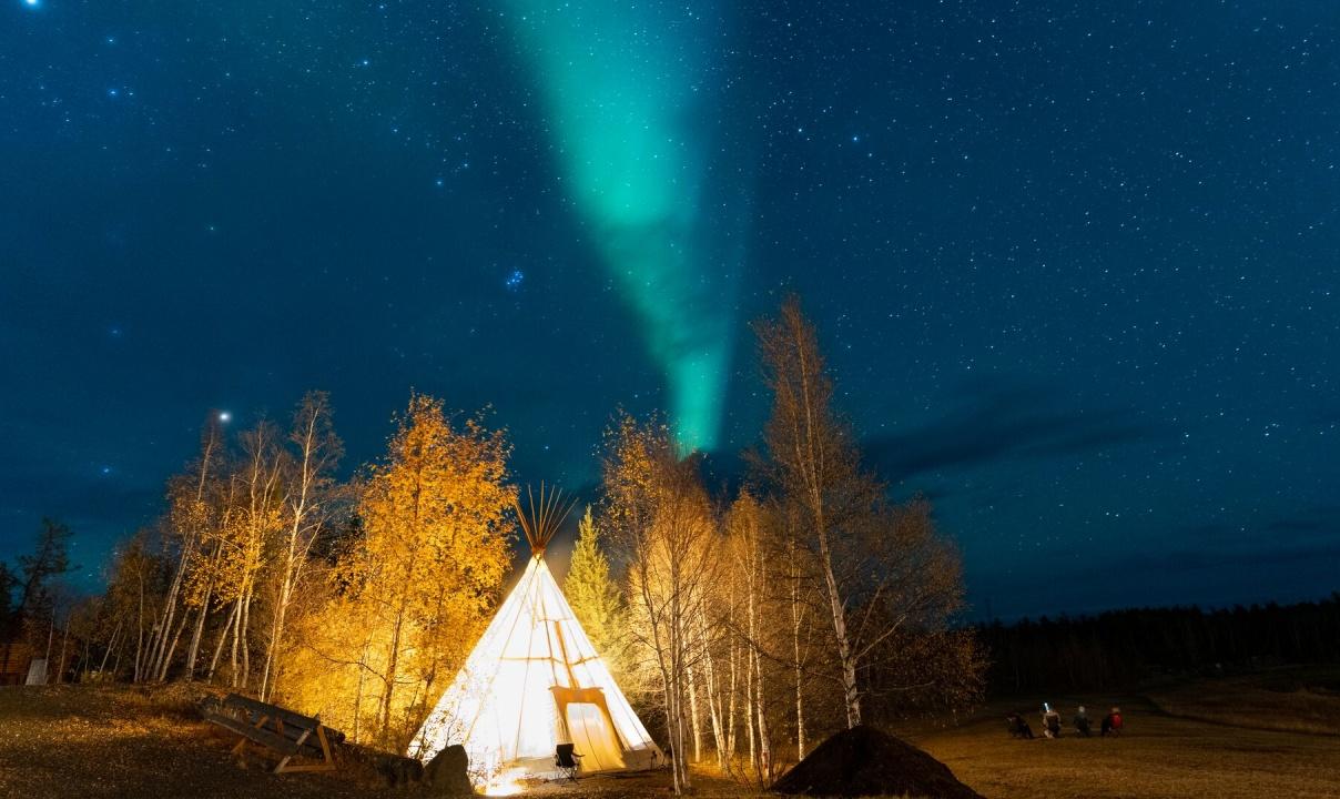 A tipi lit up from the inside in front of yellow woods at night with the aurora overhead.
