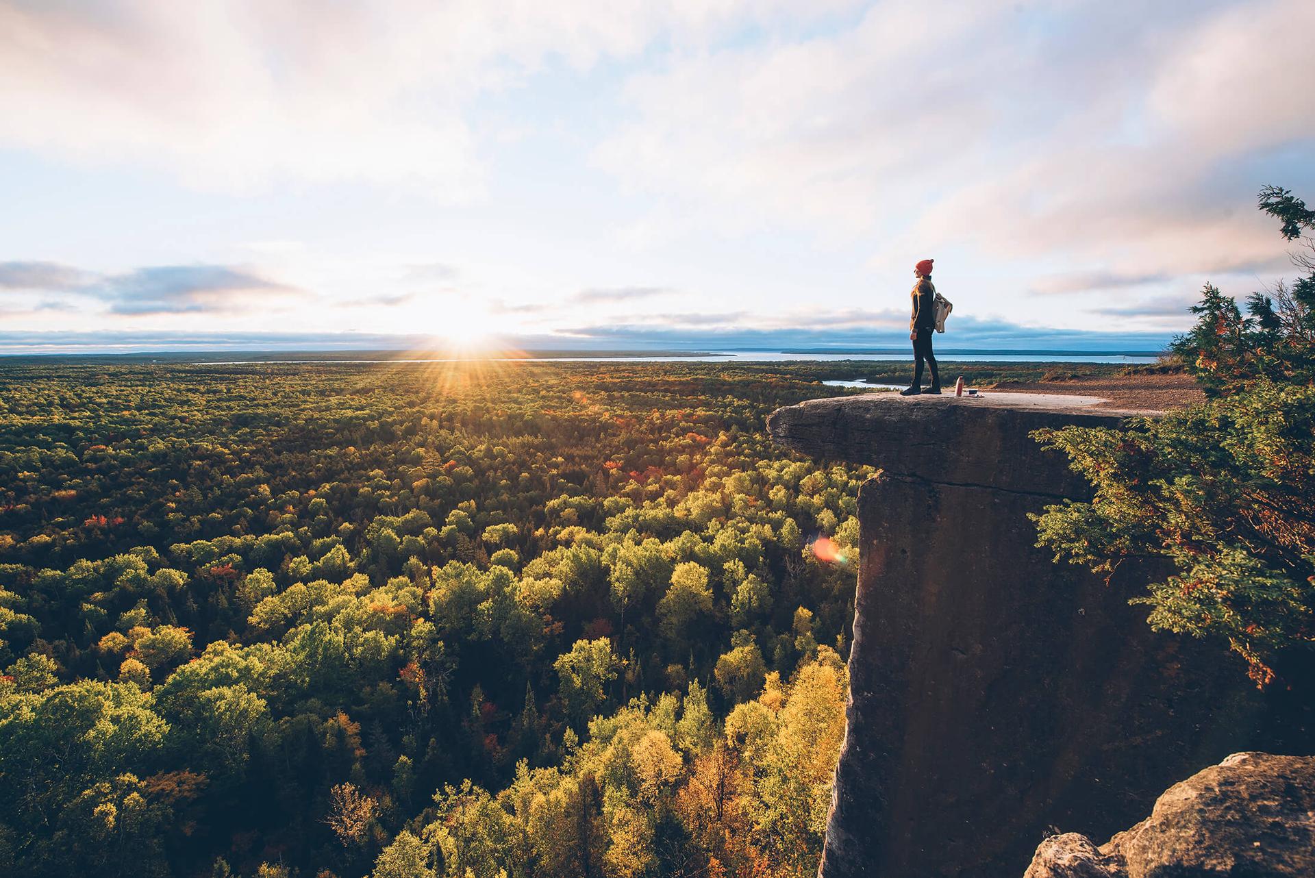 A person stands on a ridge with the sun on the horizon