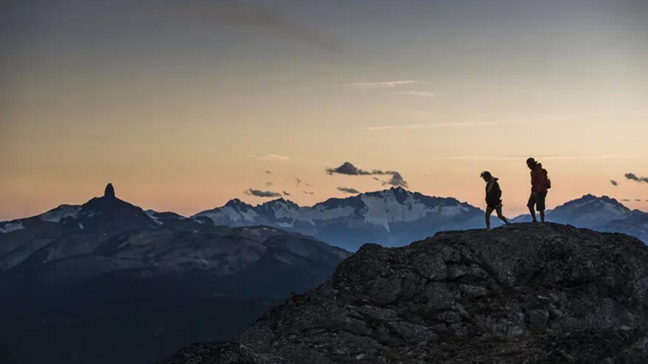 Two hikers seen in silhouette walking on a ridge in a mountain range at dusk.