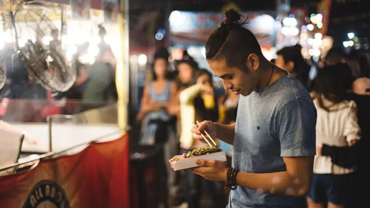 A man eating takeout food from a box with chopsticks next to a food truck at an outdoor night market.