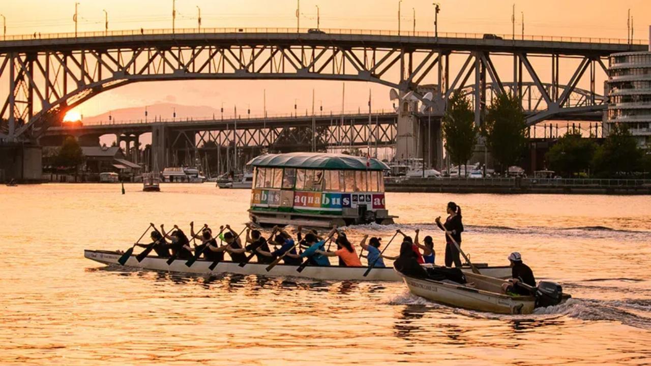 Dragon boaters paddling in front of an Aquabus in False Creek as the sun sets behind the Cambie Street and Granville Street bridges.