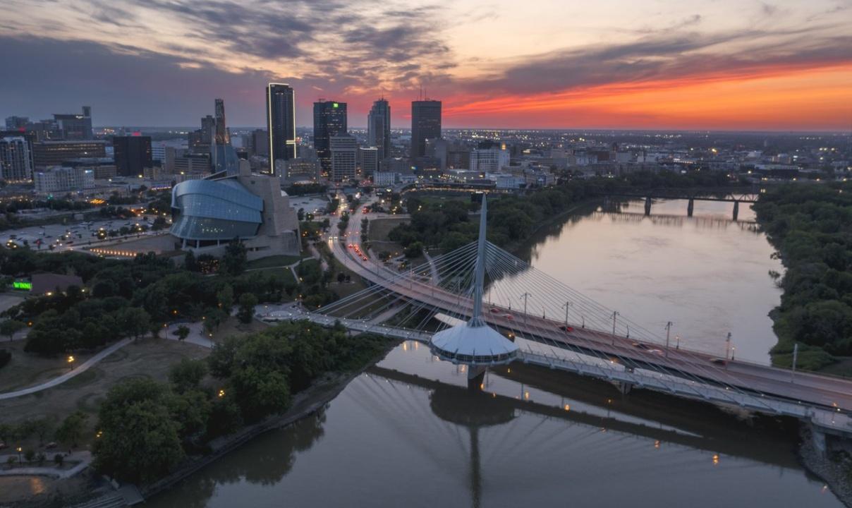 A city skyline at sunset featuring a modern suspension bridge over a calm river