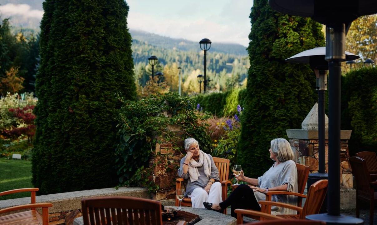 Two older women sitting on a patio with forested hills in the background, talking and drinking wine.