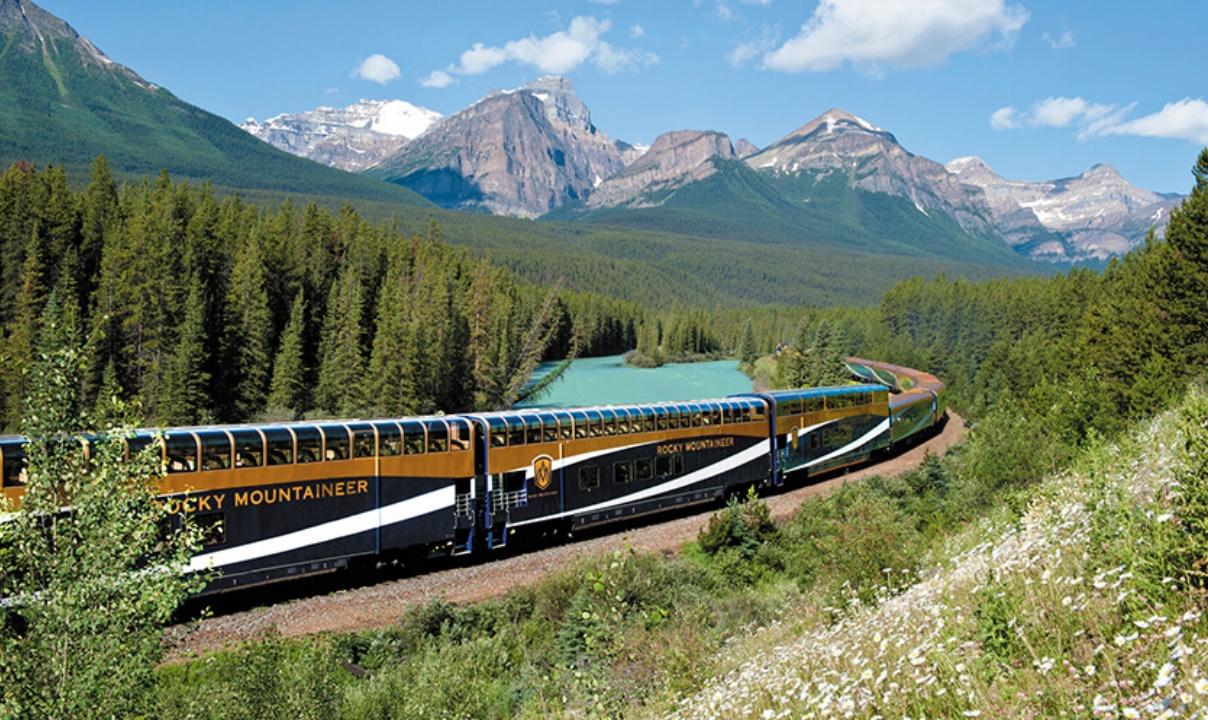 The Rocky Mountaineer passenger train passing by a river with the snowy Rocky Mountains in the background.