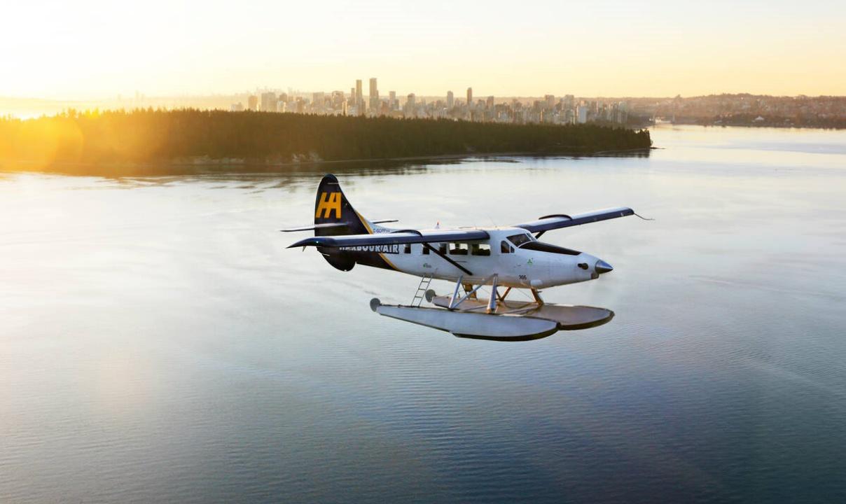 A seaplane flying over the water with Stanley Park and downtown Vancouver in the background.