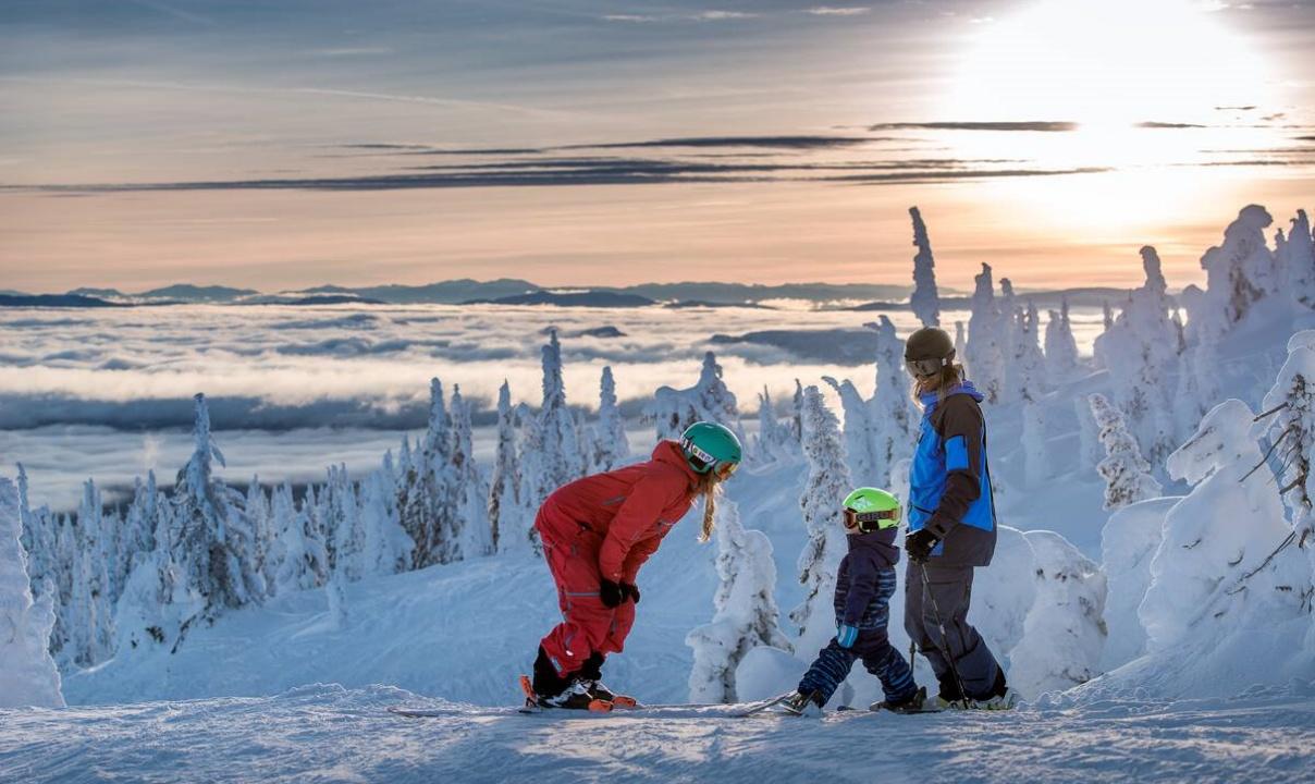 A person in a red snow suit on skis bending over to talk to a child standing next to an adult at the top of a ski run lined with snowy trees.