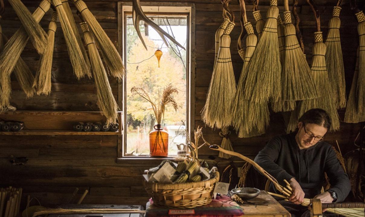 A person making a traditional broom in a workshop with multiple brooms hanging on the wall behind them.