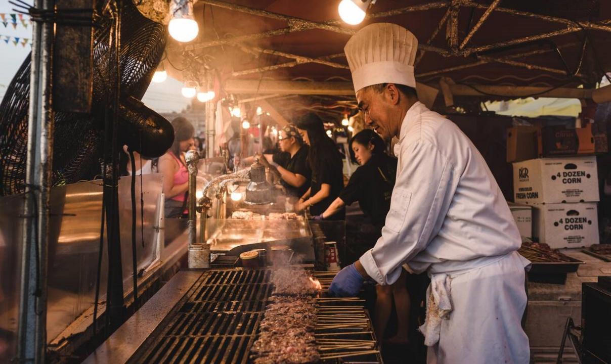 A man in a chef's toque grilling meat inside a food stall at a night market.