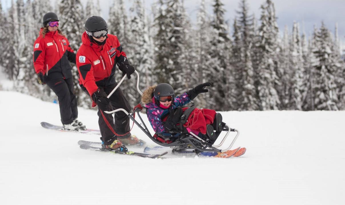 An adult on skis pushing a child on a sled while another adult looks on.