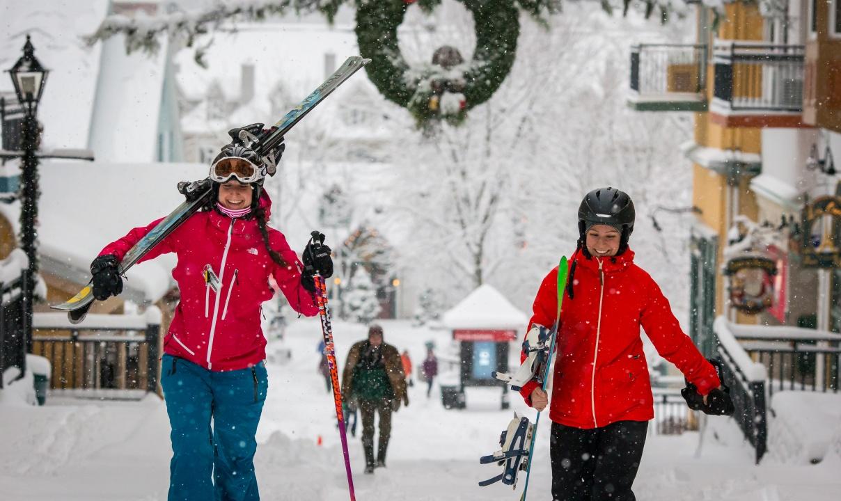 Two people in red jackets and ski helmets carrying skis on a snowy street.