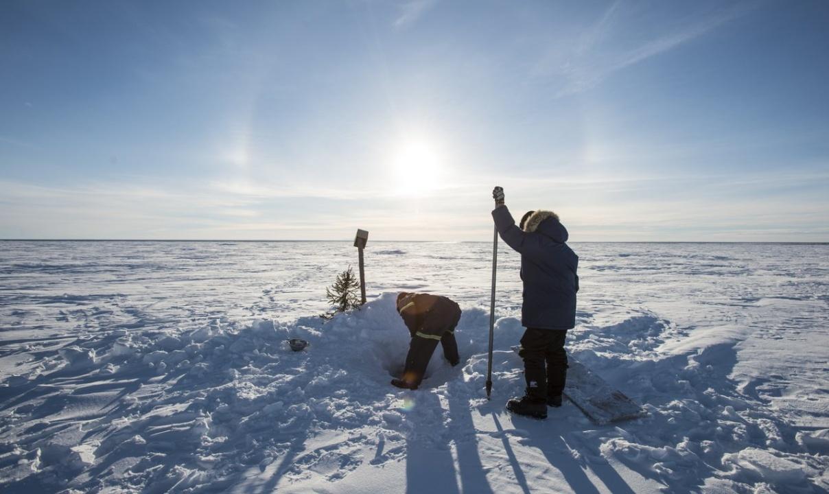 Two people digging a hole for ice fishing on a snowy, frozen lake.
