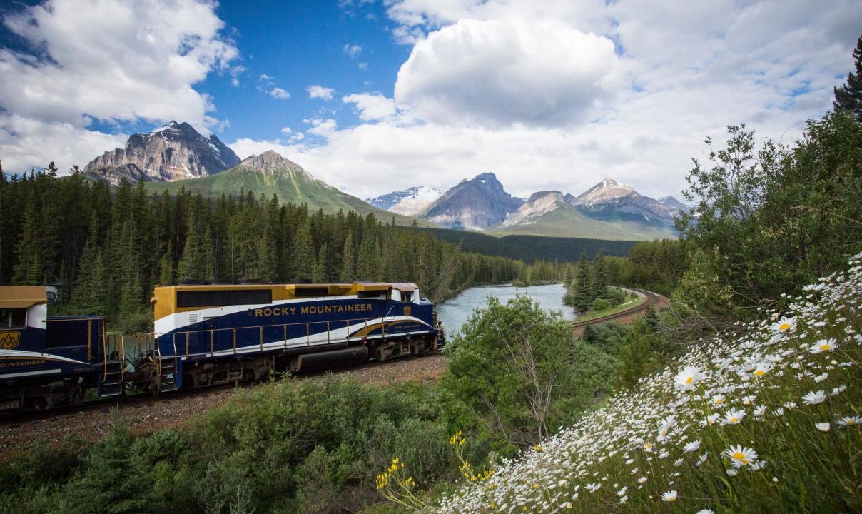 The Rocky Mountaineer train on a track by a river with wildflowers in the foreground and mountain peaks in the background.