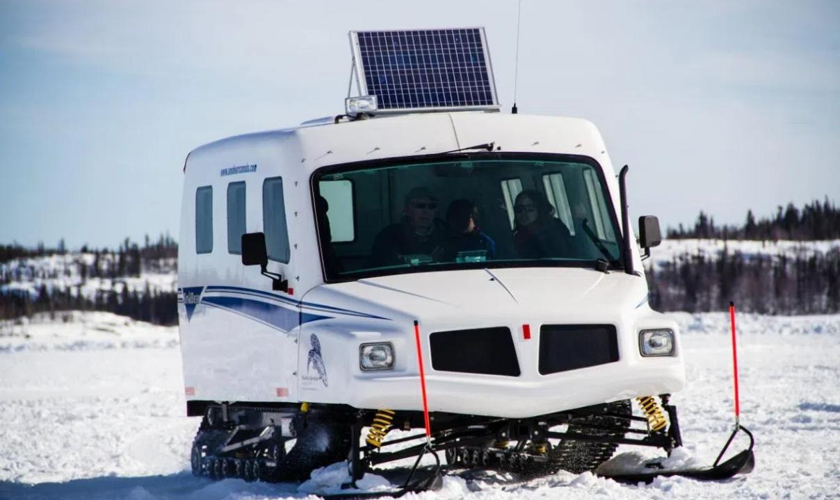 A white van on skis with a solar panel on top, in the middle of a snowy field.