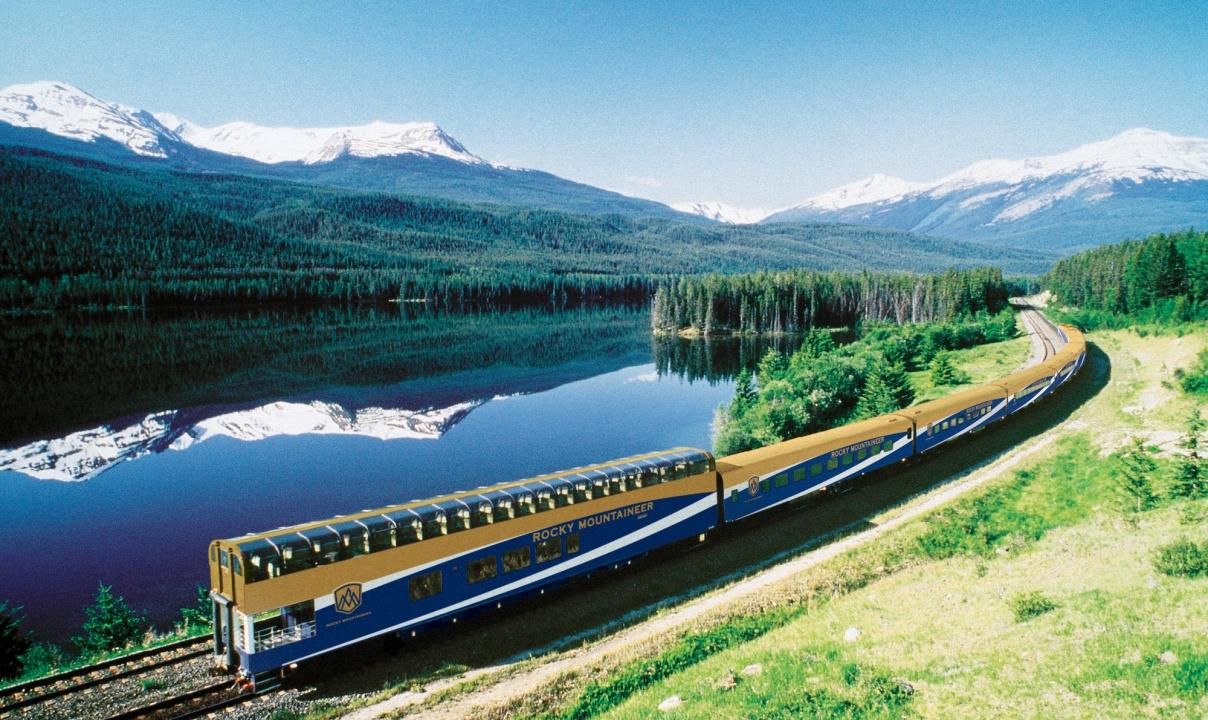 The Rocky Mountaineer train on a track by a river with snowy mountain peaks in the background.