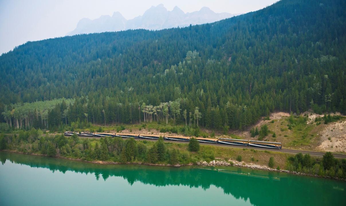 The Rocky Mountaineer train seen across a river with a forested mountain rising behind.