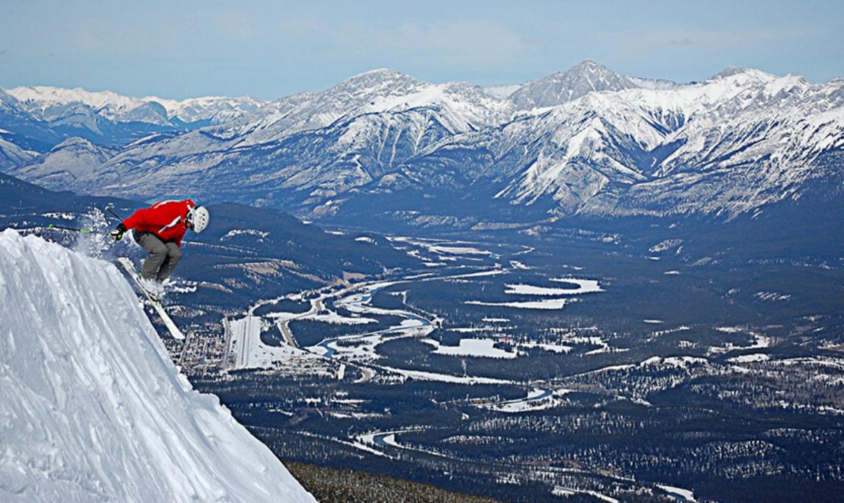 A person jumping on skis over a very steep slope with snowy mountains in the background.