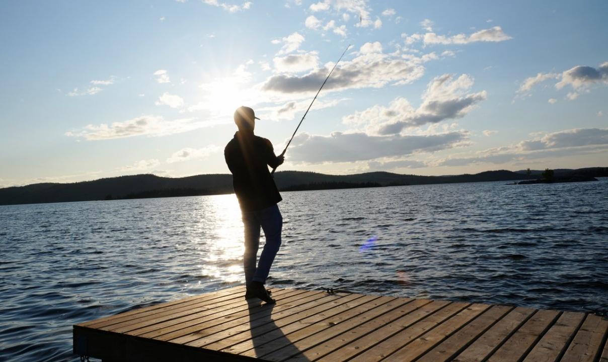 A person seen in silhouette holding a fishing rod at the end of a wooden dock.