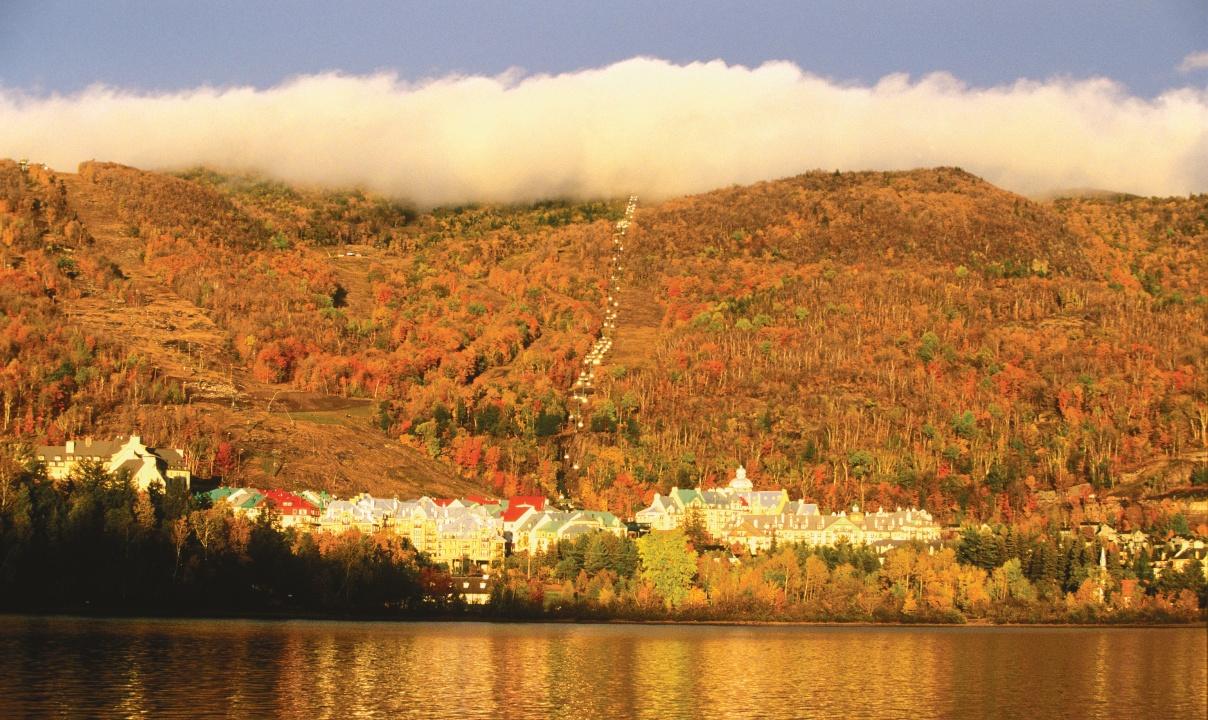 A village on the edge of a lake with an autumnal forested hillside behind.