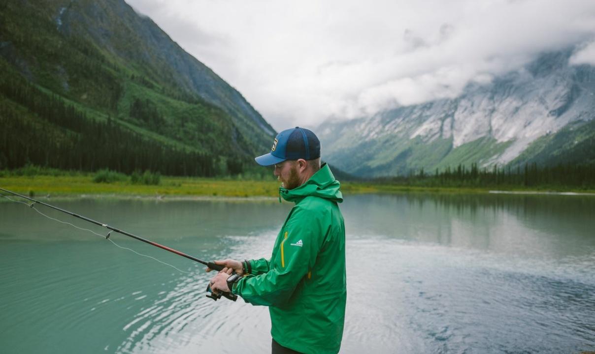 A person in a green coat fishing at a lake by mountains