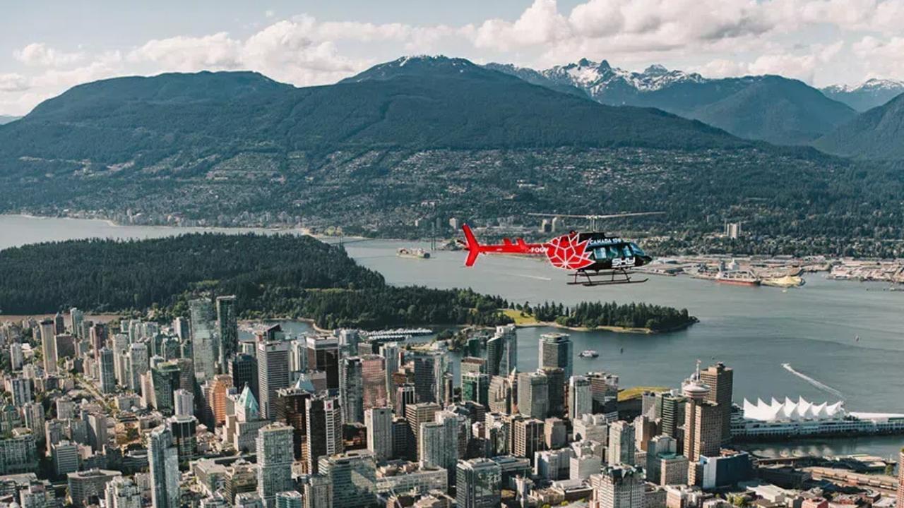 A helicopter flying over downtown Vancouver with Stanley Park and North Shore mountains in the background.