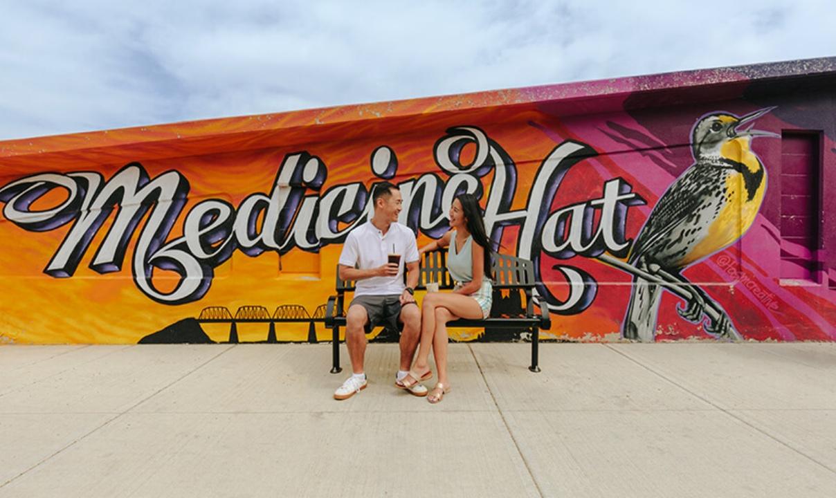 A man and a woman drinking coffee on a bench in front of a Medicine Hat mural.