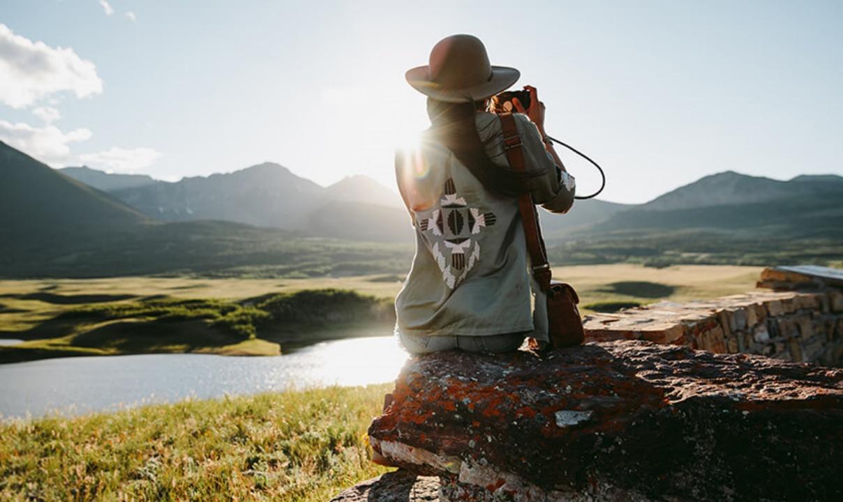A woman perched on a stone wall, her back to the camera, getting ready to take a photo of a lake and mountains in the background.