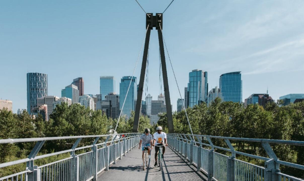 Two people riding bikes across a bridge with downtown Calgary in the background.