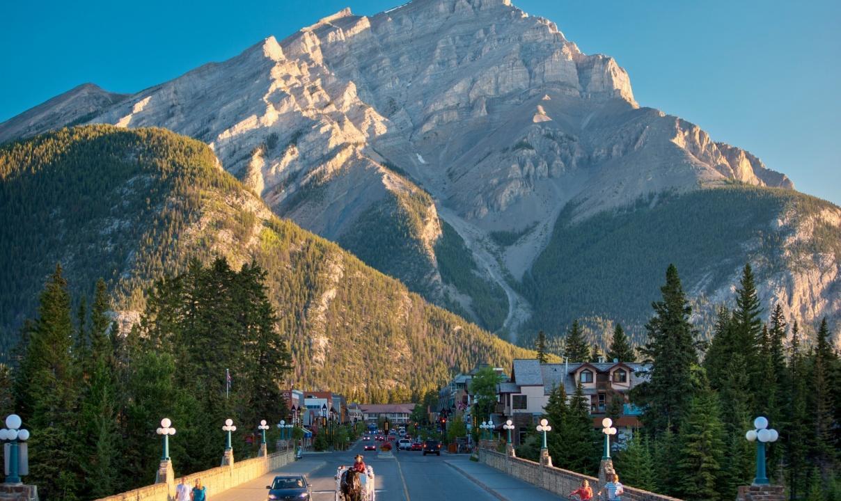 People walking and cars driving across a bridge into Banff with a large mountain looming in the background.