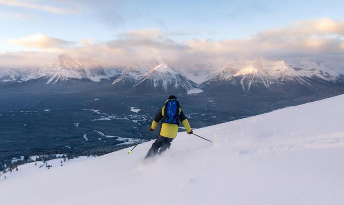 A person in a yellow and black coat and blue backpack skiing with snowy Rocky Mountain peaks in the background.