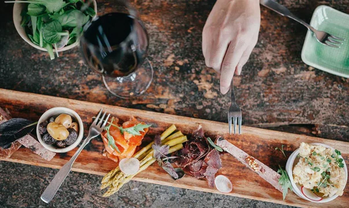 An overhead view of a charcuterie board on a table with a glass of red wine and a hand reaching towards the food with a fork.
