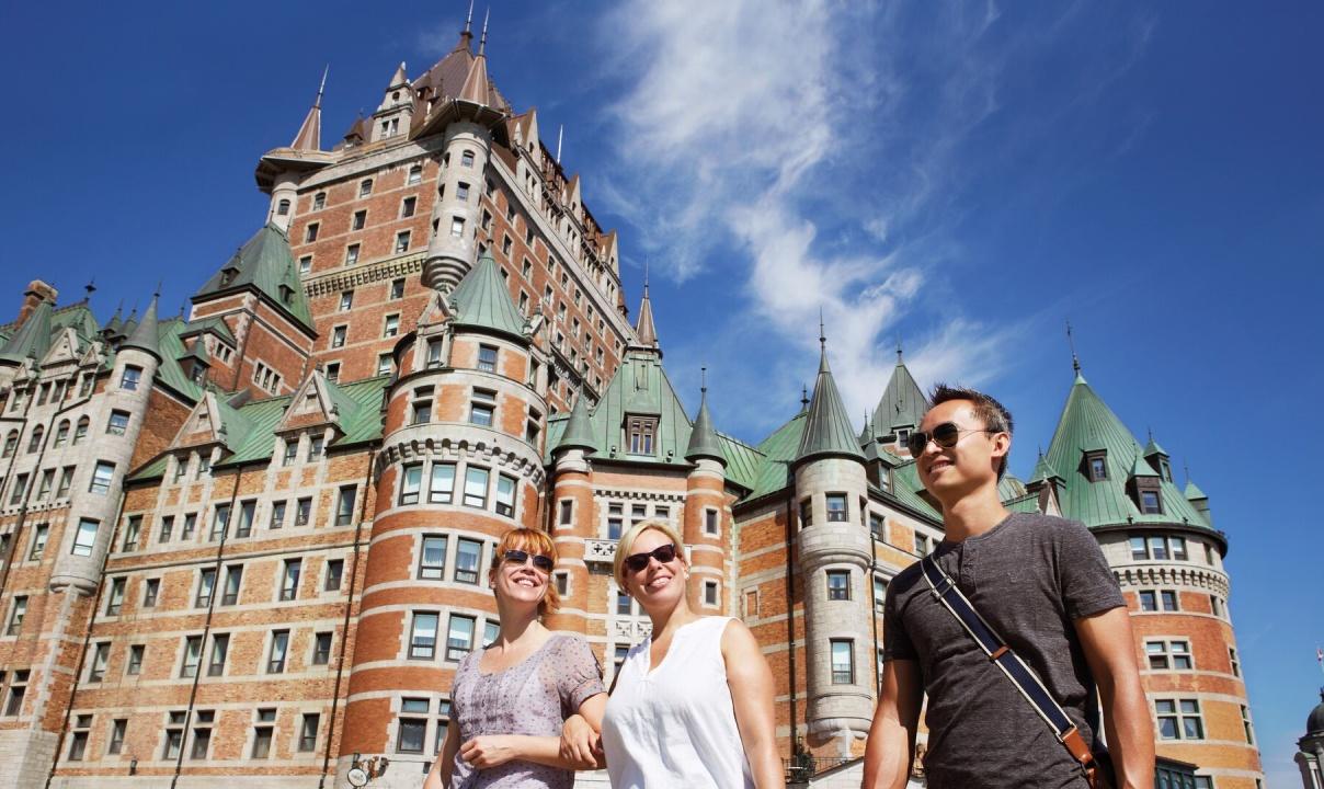 Two women and a man wearing sunglasses walking in front of the Fairmont le Chateau Frontenac on a sunny day.