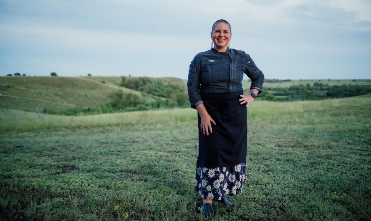 A woman standing with one hand on her hip in a grassy field with rolling hills behind.