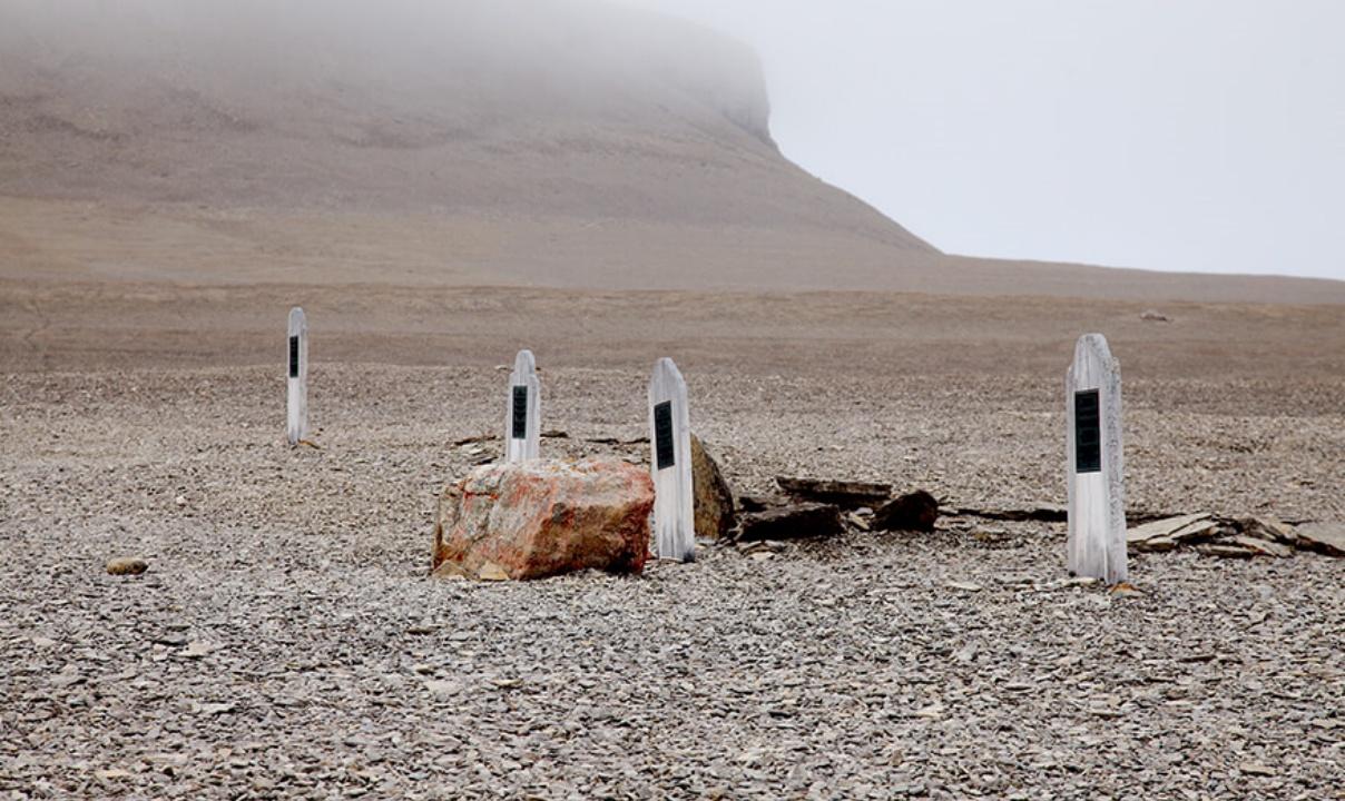 Four headstones related to the Franklin Expedition in a remote rock-filled plain on Beechey Island with a land mass rising in the background.
