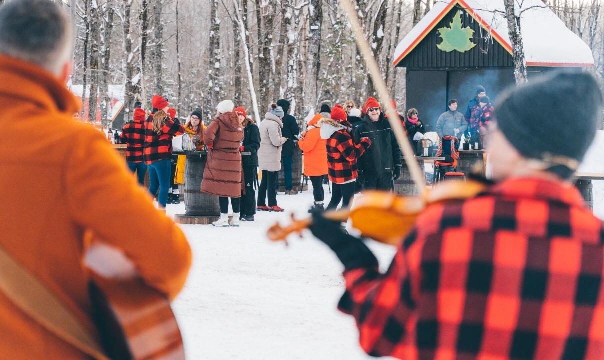 A guitarist and a fiddle player seen performing from behind as a group of people mills around a sugar shack in the snowy forest.