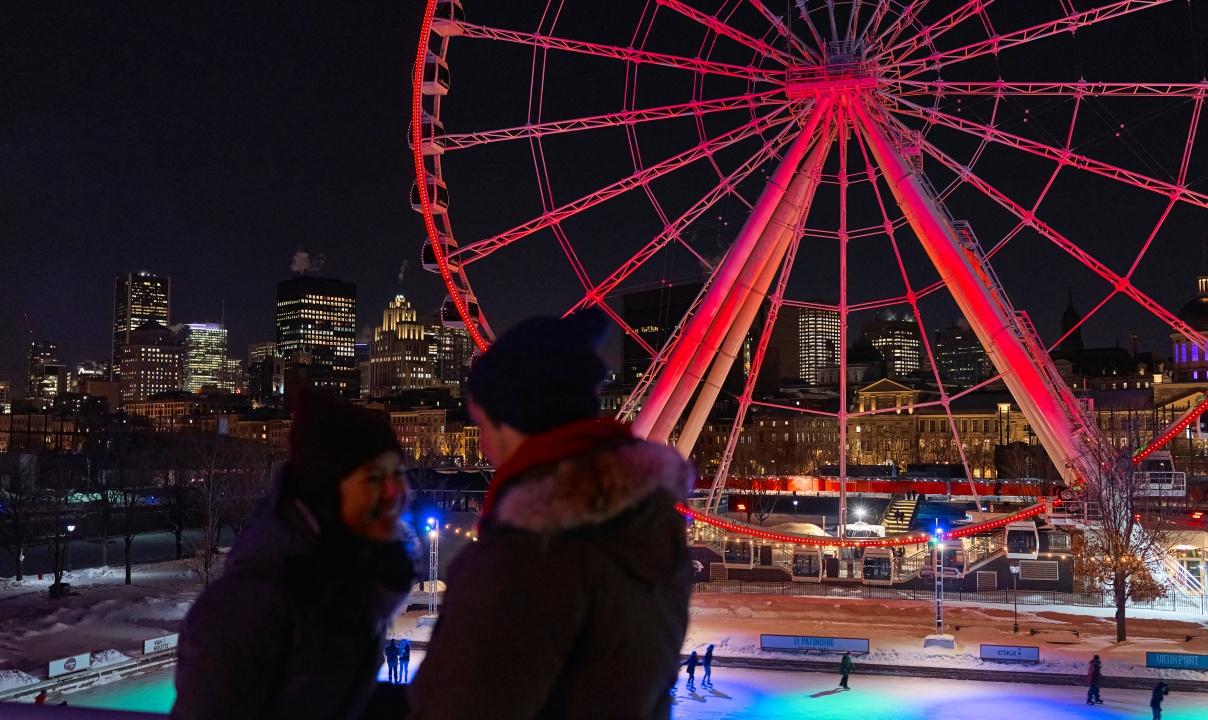 A night scene of two people talking in front of an ice rink with a red-lit ferris wheel in the background.