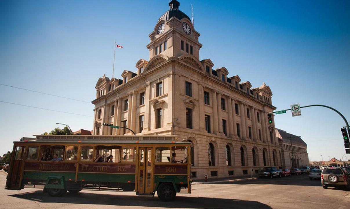 The exterior of Moose Jaw City Hall, a large building on a corner with a clock tower, and a trolley car passing in front.