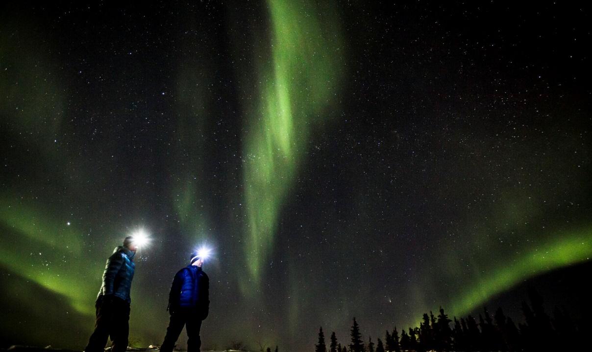Two people with headlamps turned on standing under a sky streaked with the green aurora.