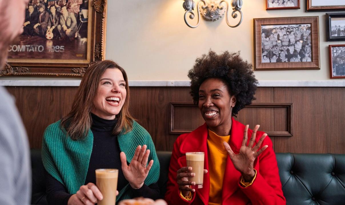 Two women drinking lattes and laughing together in a coffee shop.