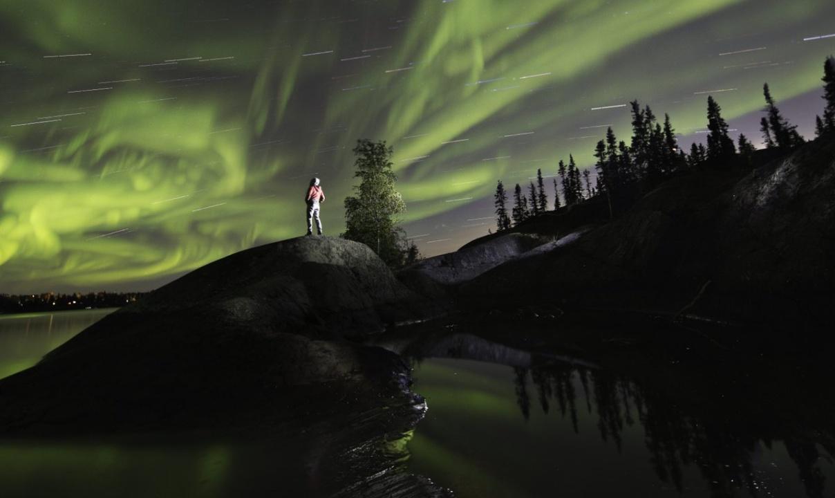 A nighttime view of a person standing on a rocky rise in the water next to a tree-lined shore, observing the sky filled with the green aurora and streaking stars.