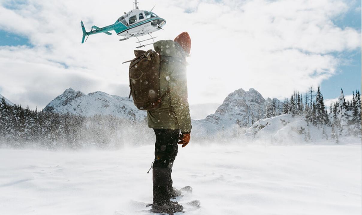 A person on skis in a snowy field surrounded by mountains and trees watching as a helicopter takes off.