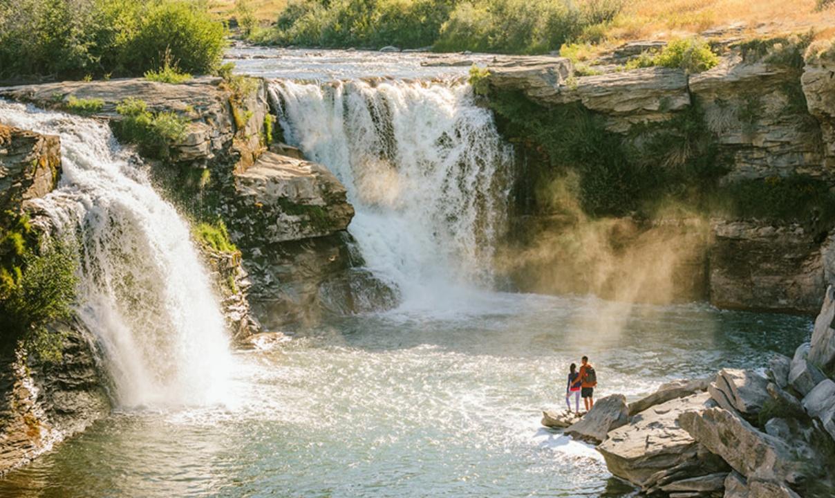 Two people standing on rocks at the edge of a pool fed by two waterfalls.