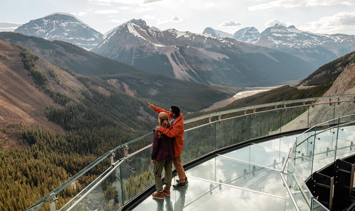 Two people standing on a glass walkway observing a scenic view of the Rocky Mountains.