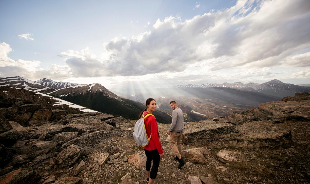 Two people hiking on top of a mountain as the sun shines through the clouds over rocky peaks in the distance.