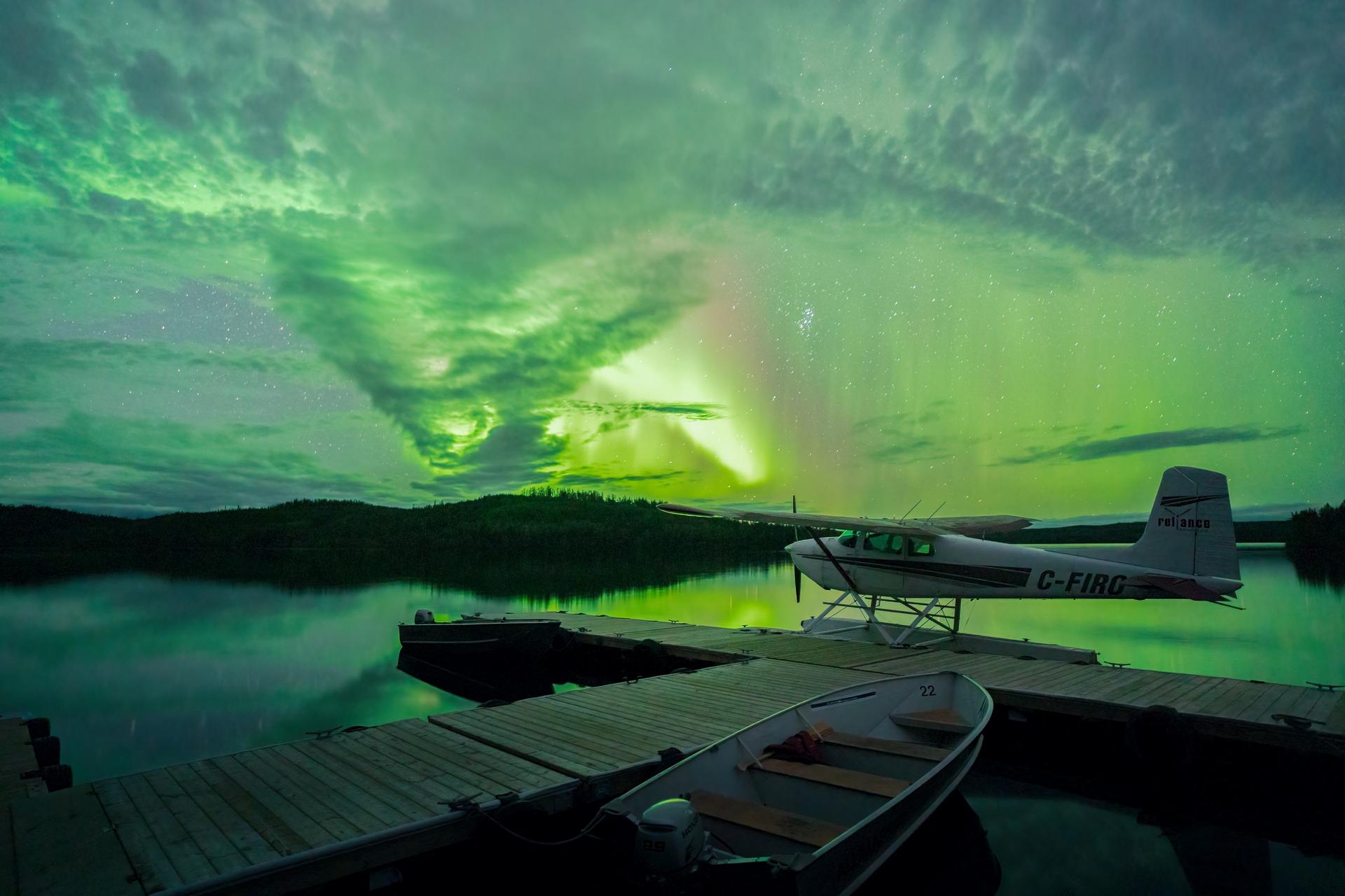 Ther northern lights over a lake with a float plane and a small boat at a dock in the foreground.
