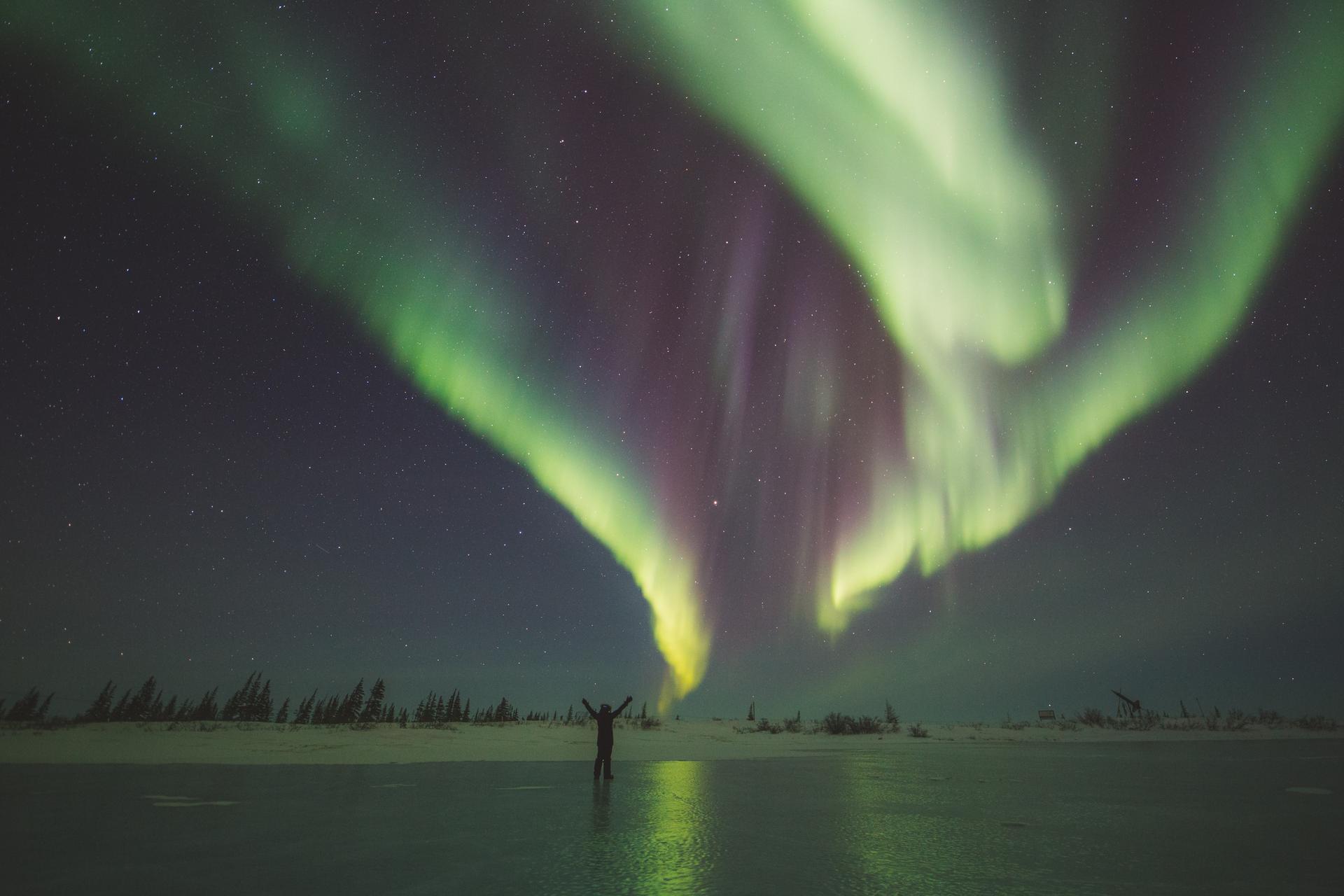 The northern lights shining above a frozen lake with a person standing in the middle with arms raised and outstretched.